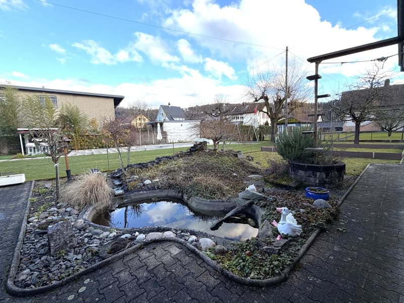 A small pond with rocks and plants, a white swan statue, a brick pathway, and a grassy area with trees and plants.