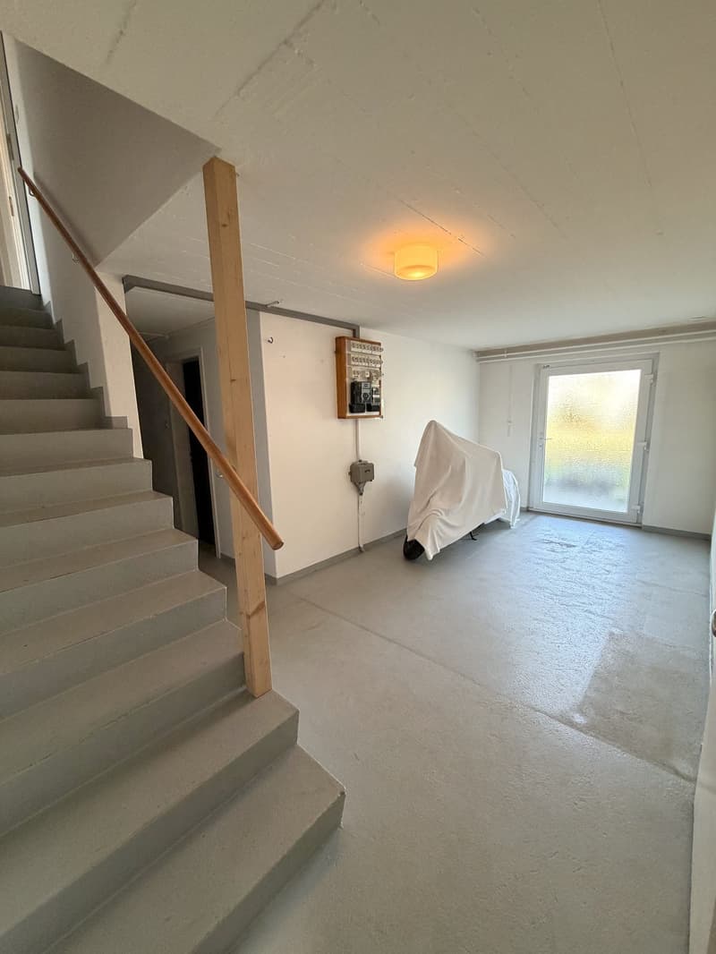 Empty room with concrete floor, white walls, wooden handrail, staircase, ceiling light, covered object, glass door, electrical panel