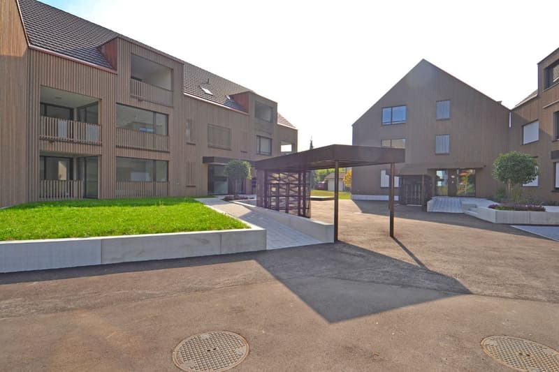 Multi-story apartment building with a modern, wooden exterior design. The building has balconies on some of the units and a covered walkway or entrance area. There is a grassy area and paved walkway in the foreground.