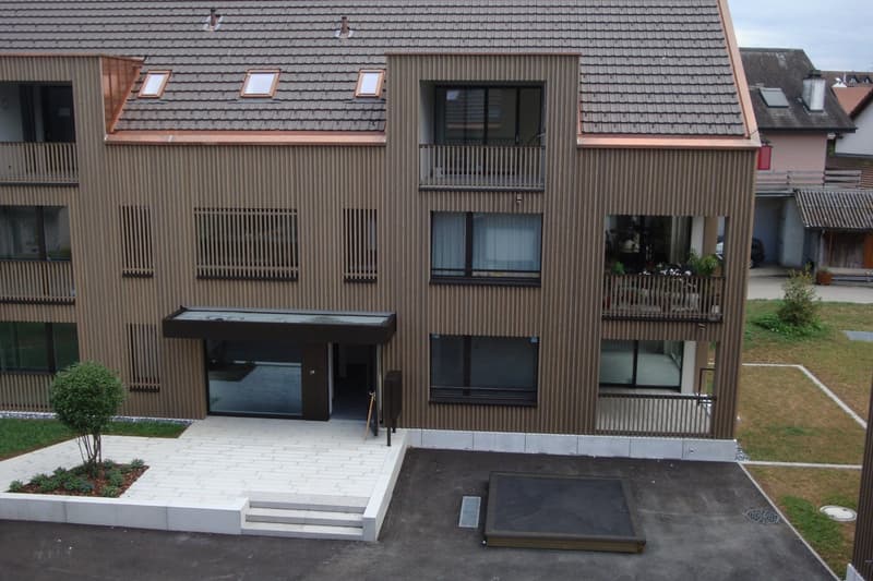 3-story apartment building with a wooden facade, balconies, and large windows. The building has a paved entrance area with steps leading up to the entrance.