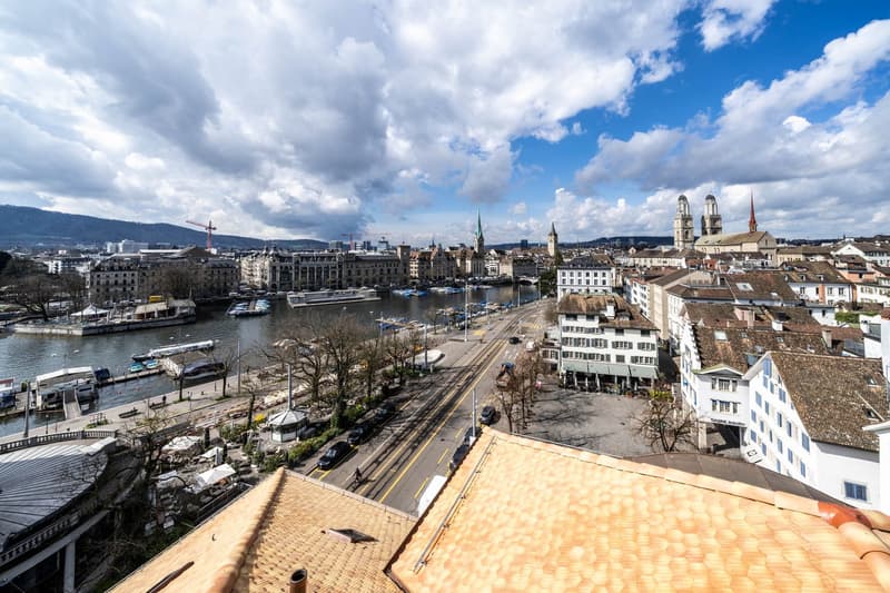 Cityscape, large river, multiple buildings, some with spires, boats docked, trees, road with cars, mountains in the distance