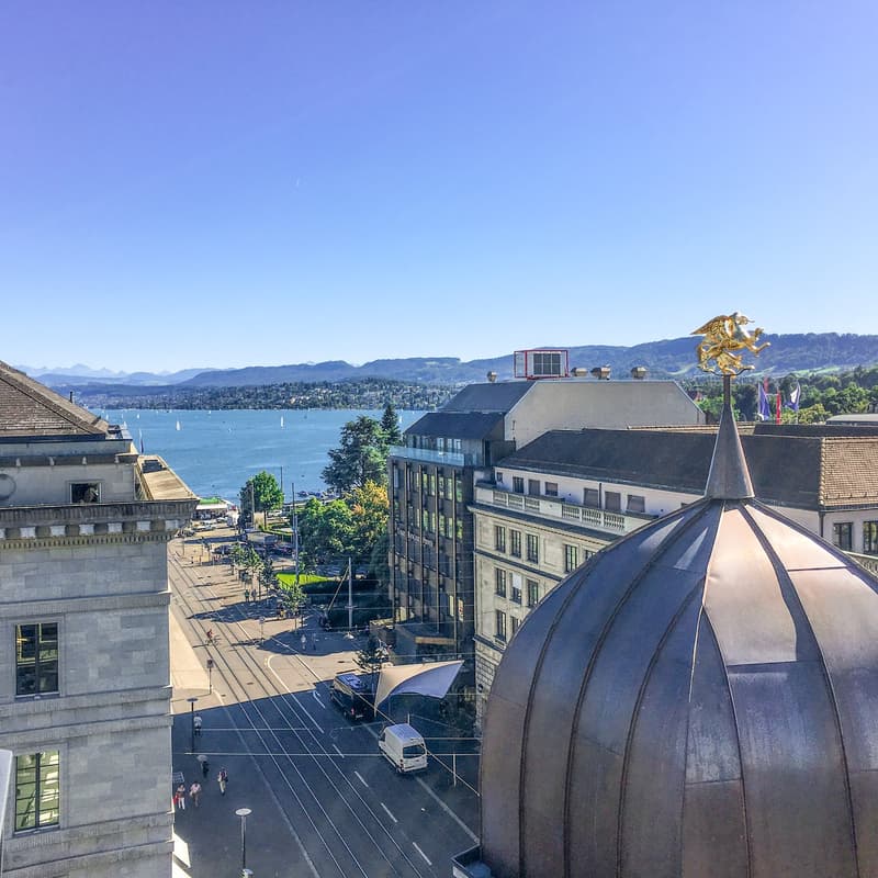 Exterior building view: Large modern buildings, golden dome on top, street with road and tram tracks, clear view of mountains and lake