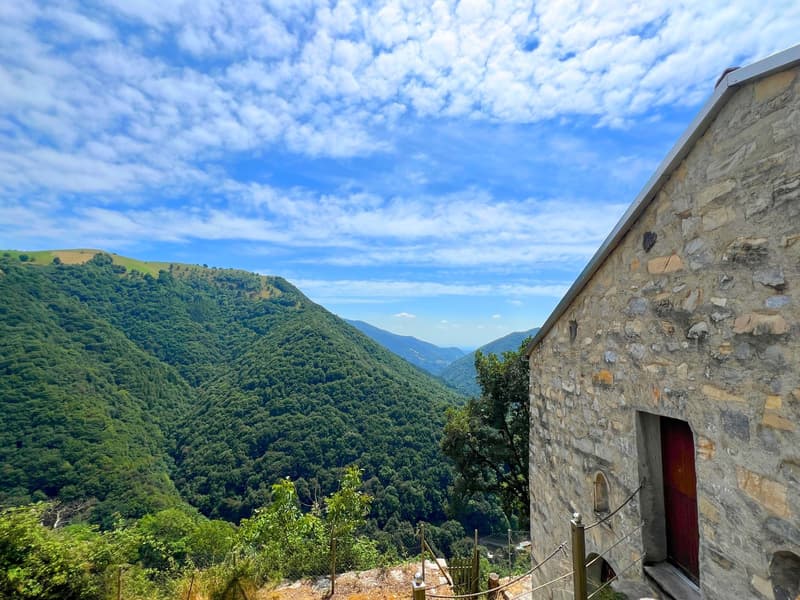 Stone building with a red door, surrounded by lush greenery, overlooking a valley with blue sky and clouds