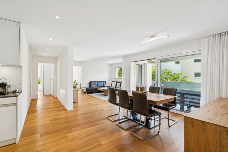 Dining area with wooden table, black chairs, glass walls leading to balcony, couch, and plant