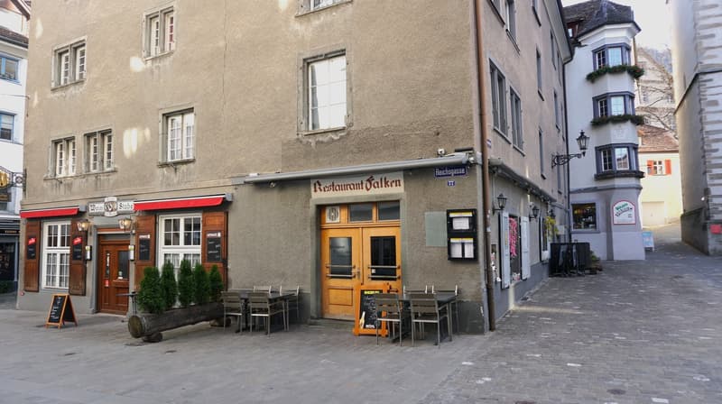 The building is multi-story with windows and a wooden door. There are plants in front of the entrance. To the right, a path leads to a narrow street with outdoor seating at a restaurant.