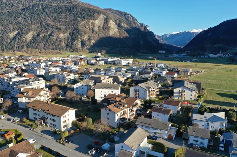 Aerial view of residential houses and apartments located in the foothills of a mountain range, featuring multi-story buildings, balconies, and trees.
