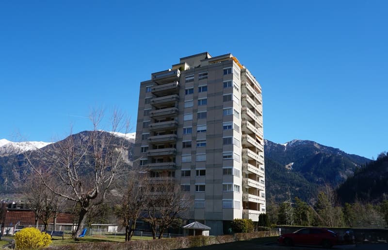 High-rise building with multiple floors and balconies, surrounded by trees and mountains