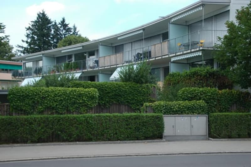 Two-story apartment building with balconies and awnings, surrounded by bushes and trees