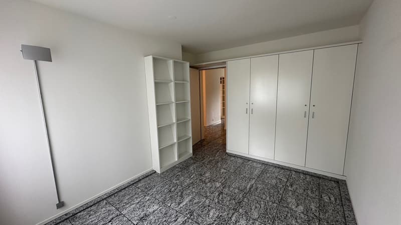 The image shows a spacious bedroom with white walls, a built-in wardrobe system along one wall, and a granite-tiled floor. The room appears to have ample storage space and a clean, minimalist design.