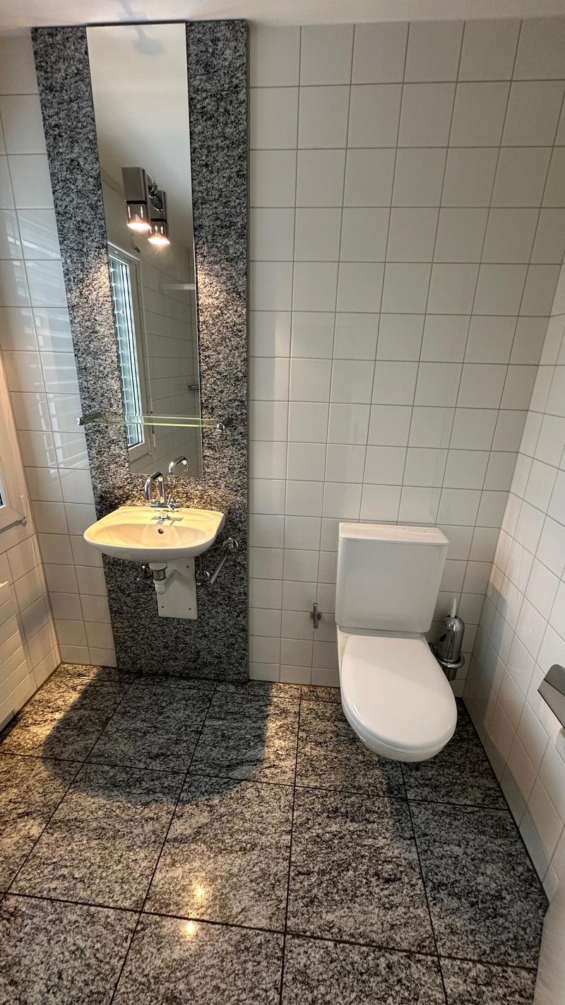 The image shows a bathroom with a granite vanity top, a white sink, a mirror, and a toilet. The floor is tiled with a granite-like pattern. The walls are tiled with white tiles.
