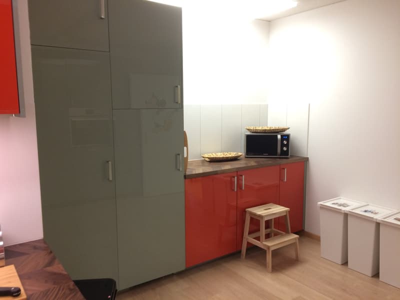 A small kitchen area with gray and red cabinets, a wooden countertop, and a microwave. Adjacent is a green tall cabinet and a wooden stool. Two storage bins are on the right side.