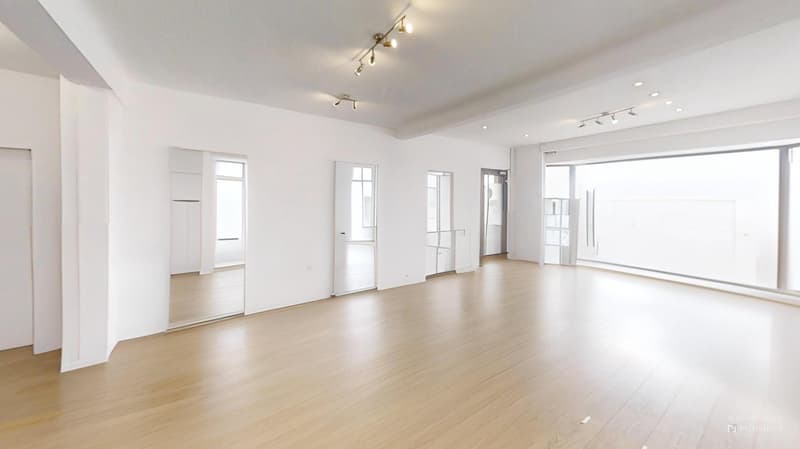 Spacious open-plan living area with white walls, hardwood floors, and recessed lighting. Multiple windows provide ample natural light. The room appears to be empty, ready for furnishing and decor.