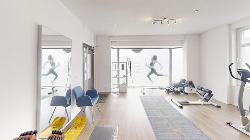 The image shows a modern and well-equipped fitness studio. The room has a bright and open layout with large windows, hardwood floors, and track lighting. There are various fitness equipment such as treadmills, exercise machines, and free weights. The wall