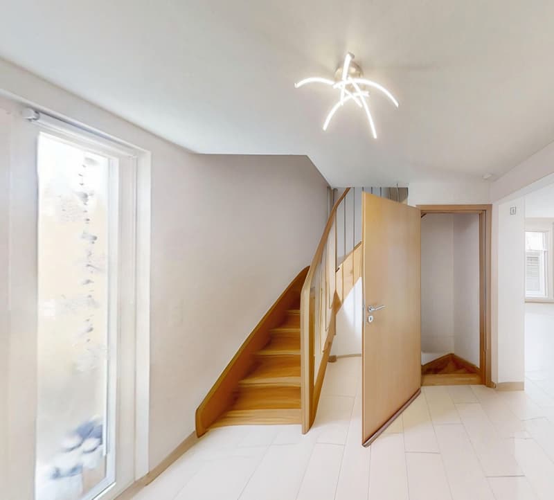 An empty hallway with white tiled floor, a large open door, wooden spiral staircase, large window with frosted glass, white walls, ceiling with modern light fixture.