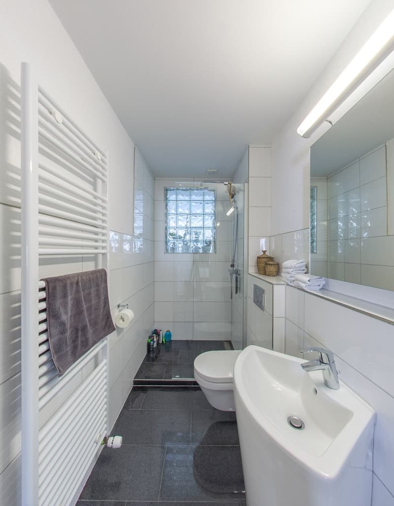 modern bathroom with white tiles, white sink, white toilet, glass shower enclosure, towel rack, mirror, towel