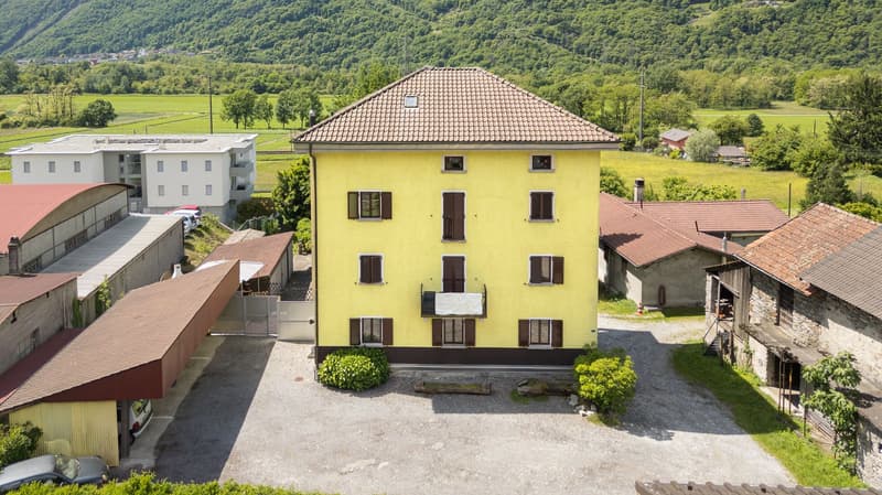 Multi-story yellow building, brown roof, several windows, surrounded by greenery, neighboring buildings, fenced yard, cars parked in front, distant mountain view.