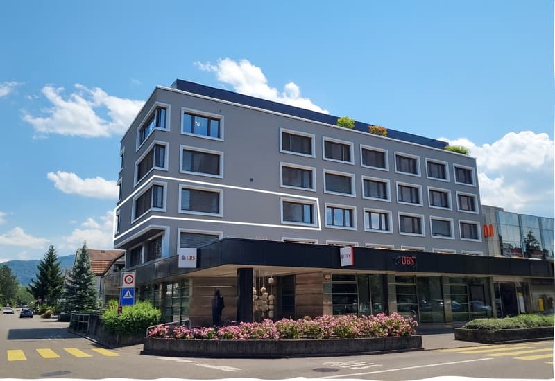 gray multi-story building with a raised ground floor, white-framed windows, plants on the roof, signage of UBS