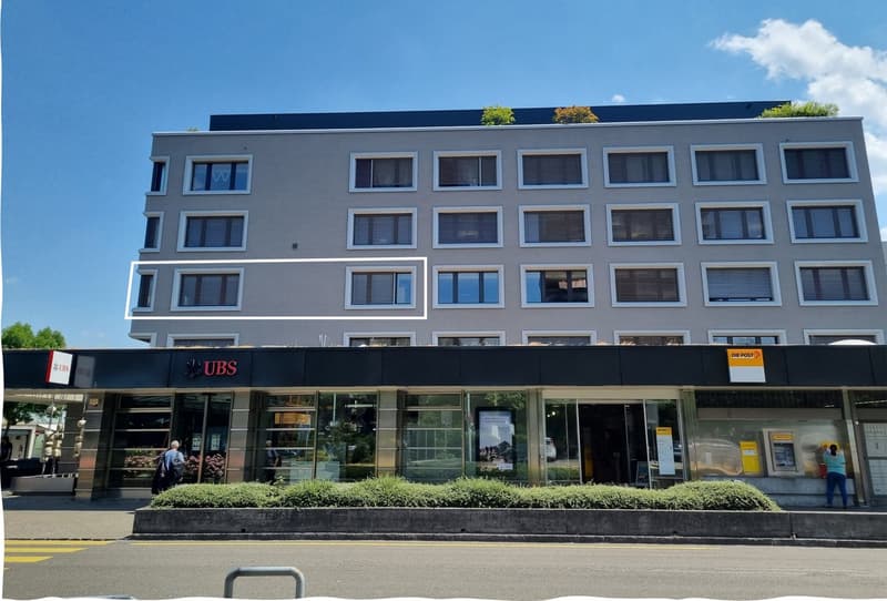 3 story apartment building, gray exterior, many windows, entrance with UBS sign, green plants on the rooftop, UBS and Die Post signboards on the lower floor