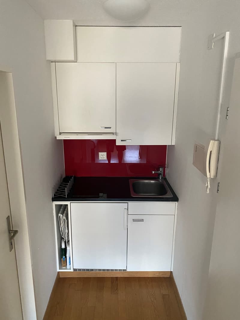 modern kitchen with white cabinets, red backsplash, black countertop, stainless steel sink, faucet, electrical outlet, and a small open cabinet with a washing machine