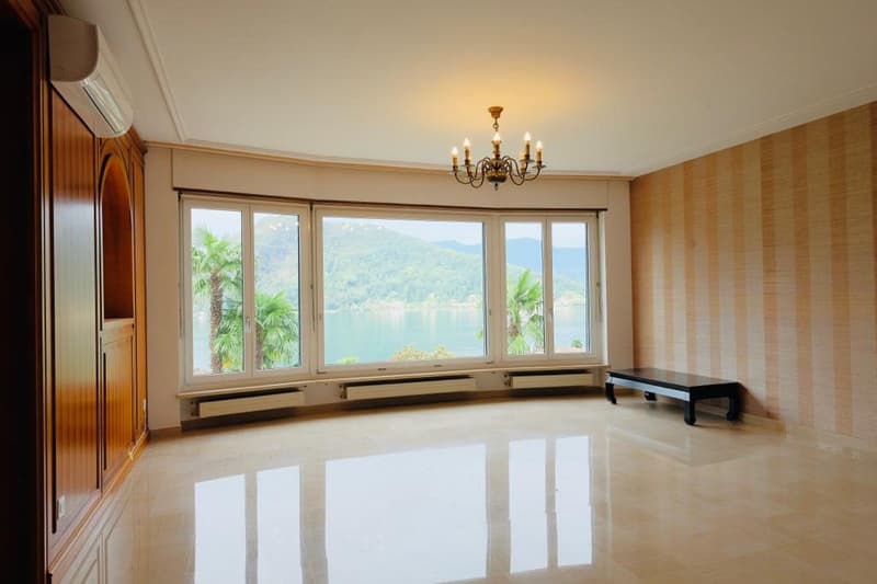 Spacious living room with large windows offering a scenic view of the mountains and palm trees. The room features a wooden cabinet, a chandelier, and a raised platform with a bench-like seating area.
