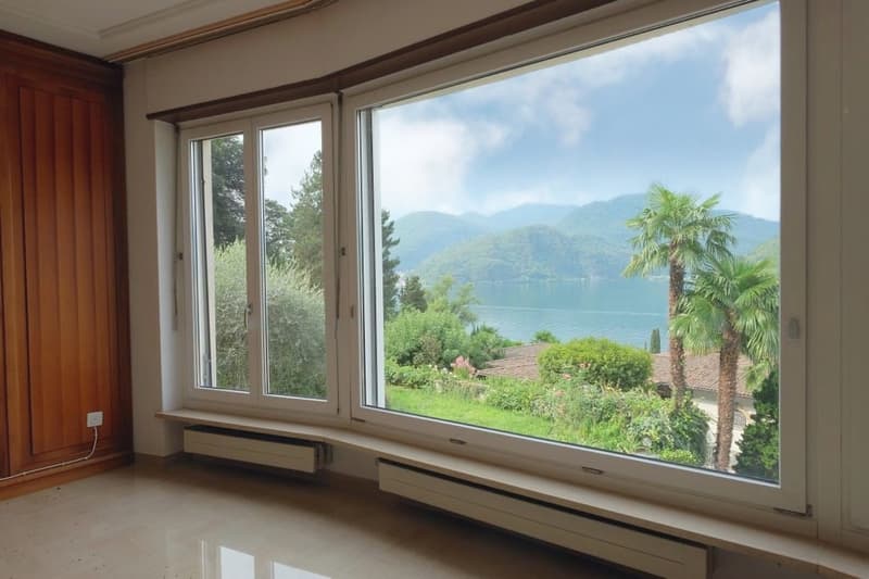 Large windows with a panoramic view of a lake surrounded by mountains and lush greenery, including palm trees. The room has wooden paneled walls and a tiled floor.