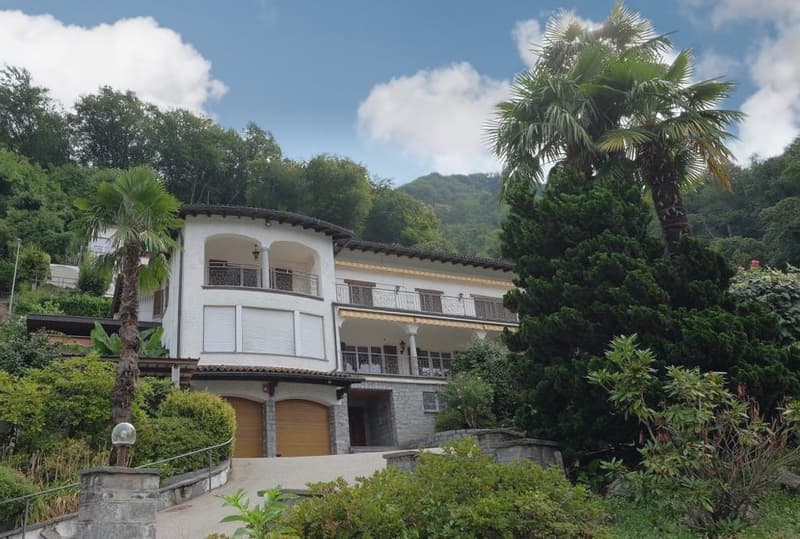Two-story white villa with arched windows, balconies, and a tiled roof surrounded by lush greenery and palm trees