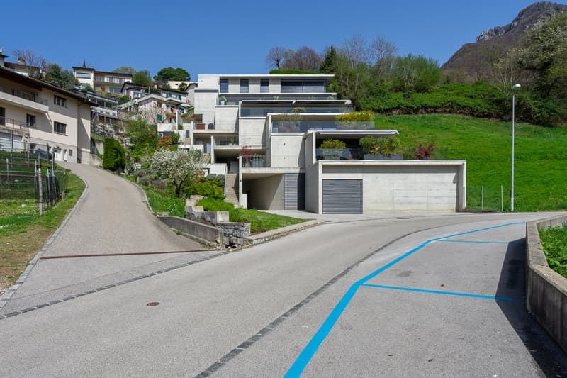 The image shows a residential area with modern multi-story apartment buildings nestled into a hillside. The buildings have a clean, minimalist design with large windows and balconies. There is a paved road with a blue line marking running through the scen