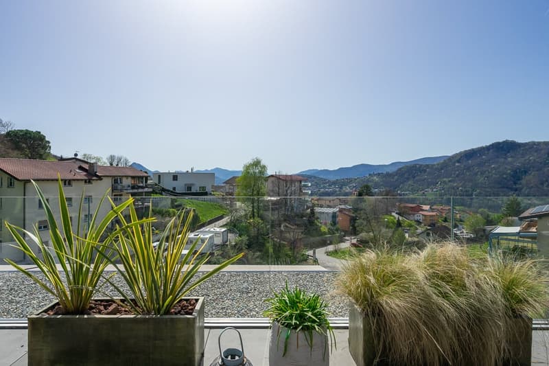 The image shows a balcony or terrace overlooking a residential area with houses, buildings, and mountains in the background. The balcony has potted plants and greenery, and the view includes a mix of buildings, trees, and natural scenery.