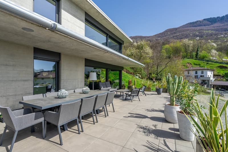 Modern concrete house with large windows, outdoor patio with dining table and lounge chairs, surrounded by lush greenery and mountains in the background