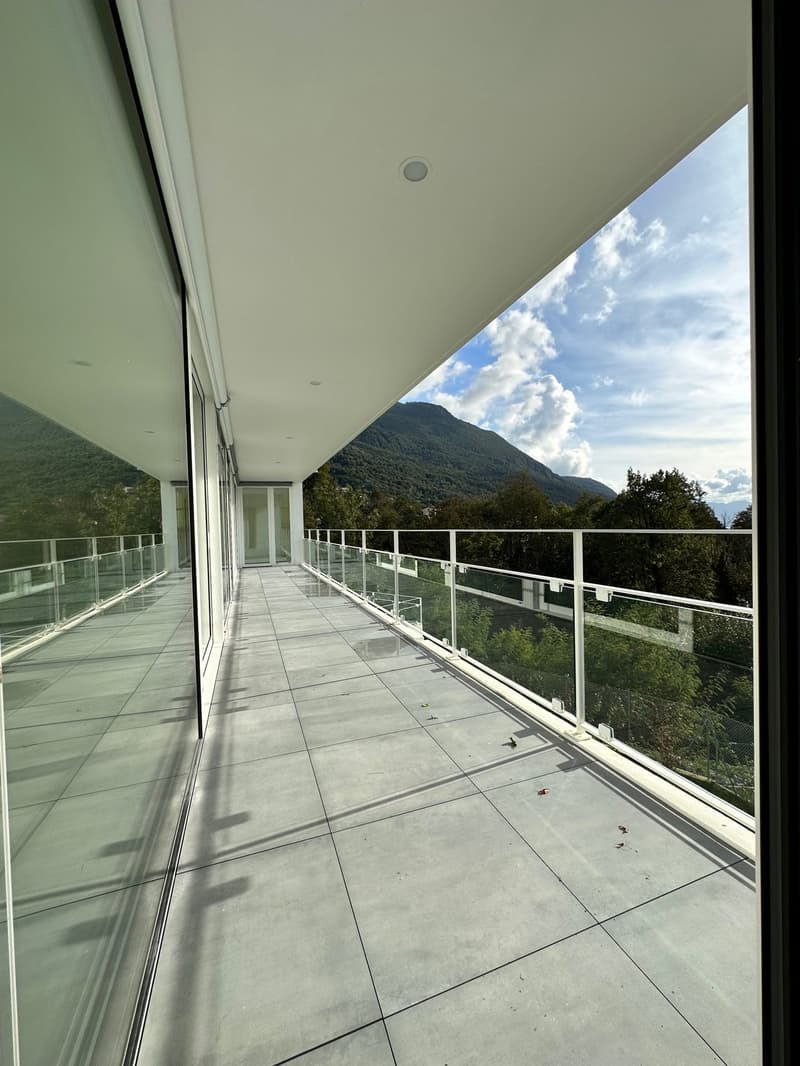 Large balcony with glass railing, overlooking a mountainous landscape with trees and cloudy sky