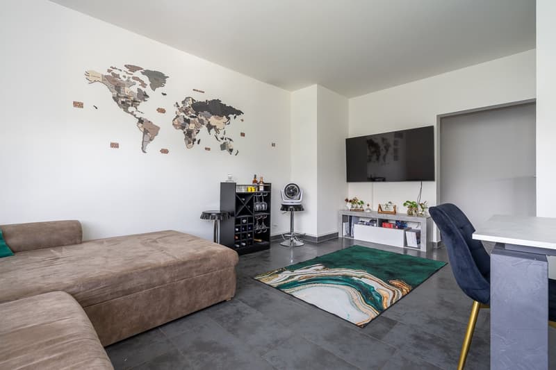 Modern living room with a large world map wall decor, a gray sectional sofa, a black entertainment unit with shelves and a TV, a green and white patterned rug, and a blue accent chair.