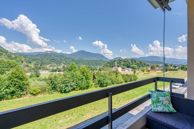 Balcony overlooking a scenic landscape with mountains, forests, and a village in the distance. The balcony has a black railing and a decorative tropical-themed pillow.