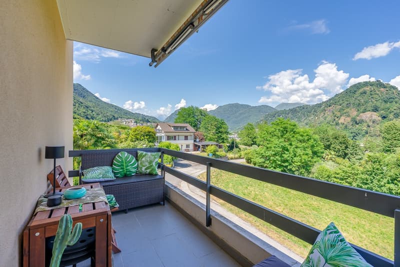 Spacious balcony with a scenic view of the surrounding mountains and lush greenery. The balcony features a wicker bench with tropical-themed cushions, a wooden side table, and potted plants, creating a cozy and relaxing outdoor space.
