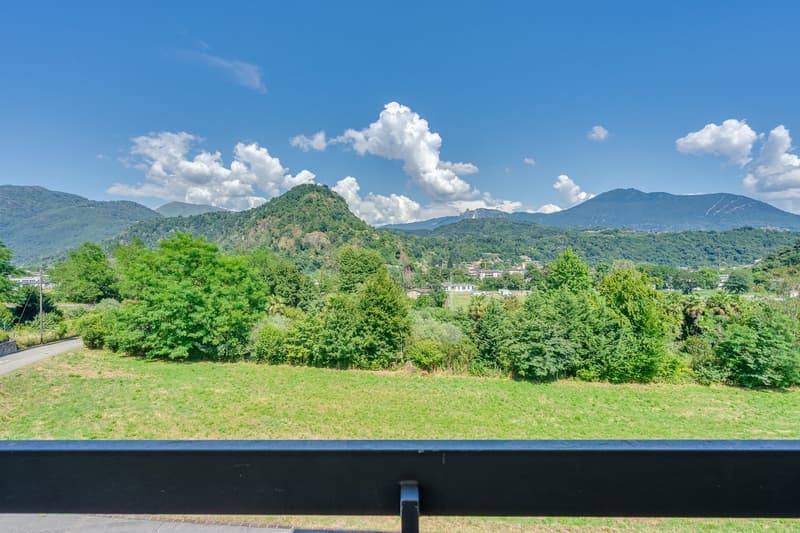 The image shows a scenic landscape with lush green hills, mountains, and a clear blue sky with fluffy white clouds. The foreground features a grassy field, and the background is filled with dense forests and towering peaks. The overall scene appears to be