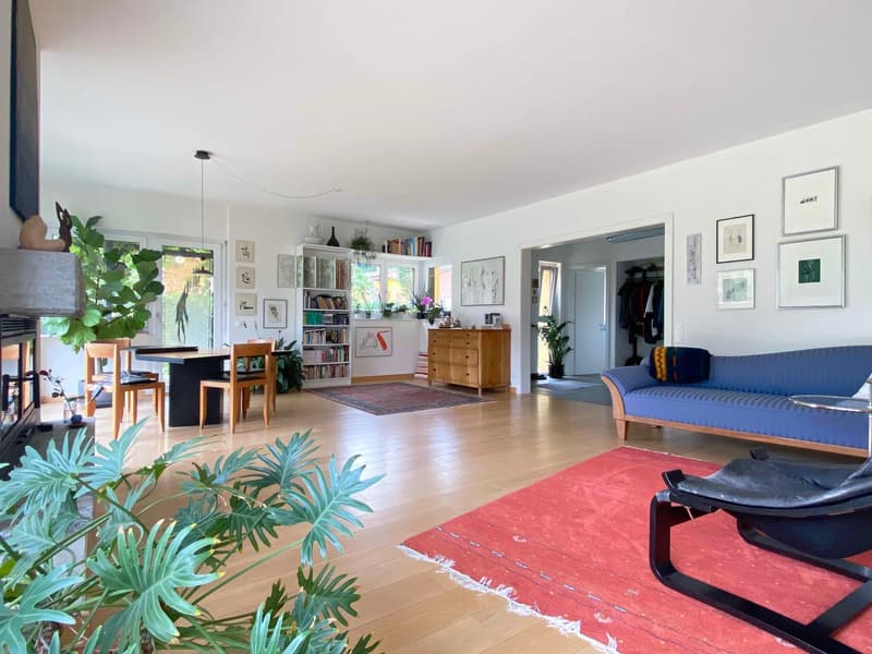 Spacious open-plan living area with hardwood floors, large windows, built-in bookshelves, potted plants, blue sofa, wooden dining table and chairs, and various artwork and decor on the walls.
