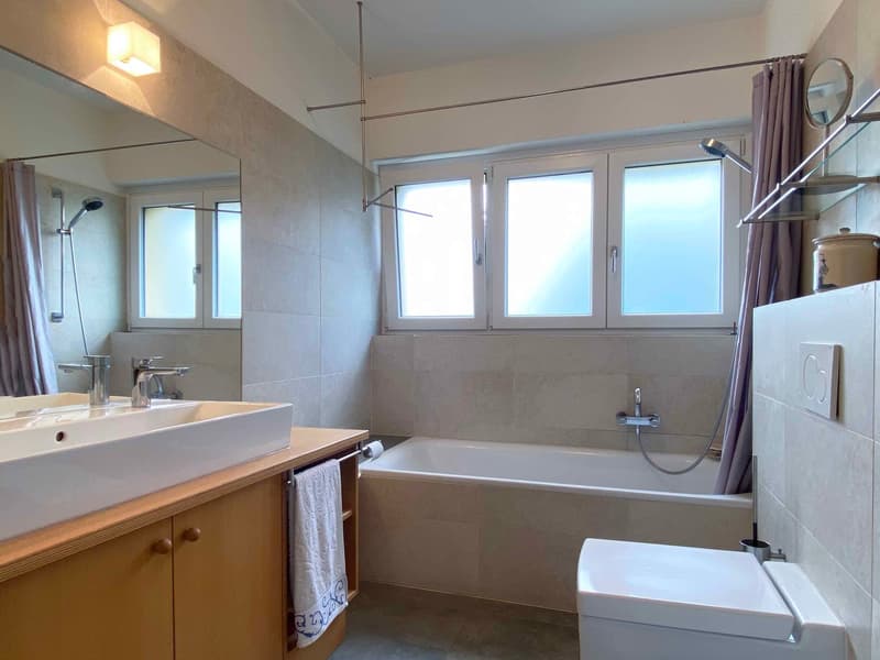 Spacious bathroom with large windows, tiled walls, bathtub, vanity sink, and a private washing machine