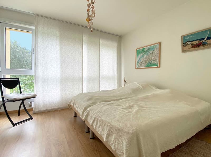 Spacious bedroom with large windows, white walls, hardwood flooring, a bed with a light-colored bedspread, and a framed artwork on the wall.
