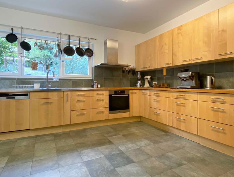 Spacious kitchen with wooden cabinets, gray tile flooring, large windows, and hanging pots and pans. The kitchen includes a dishwasher and various kitchen appliances.