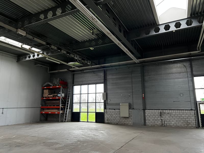 Empty warehouse with high ceiling, metal beams, large windows, storage shelves, ladder, and a door.