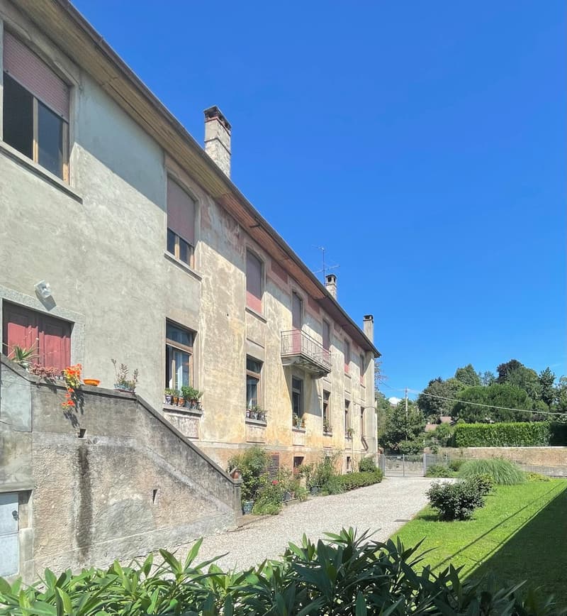Old building, multiple windows, balcony, plants, gravel driveway, garden