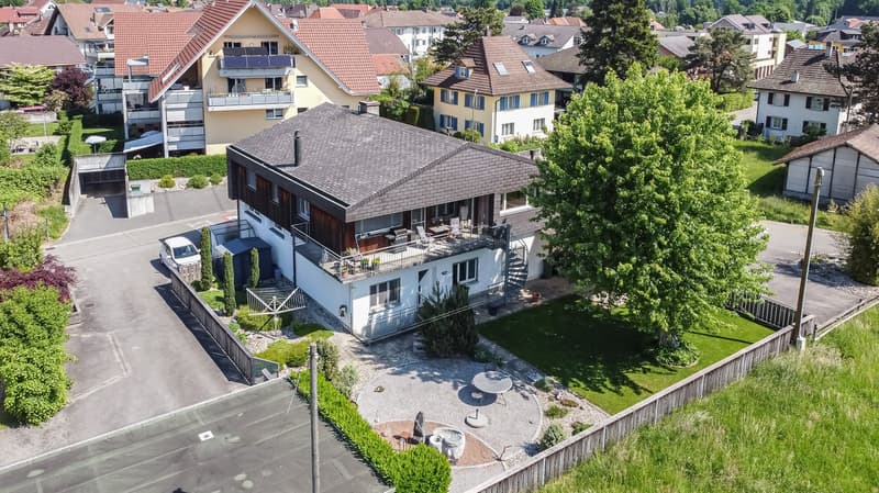 2-story house with red tile roof, wooden exterior, balconies, surrounded by trees and greenery, with a paved driveway and parking area in the front