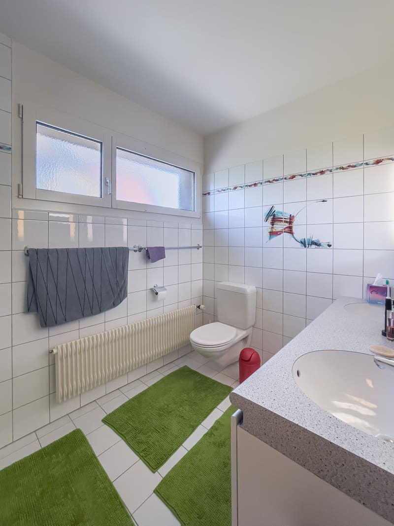 Tiled bathroom with a toilet, sink, and radiator. The bathroom has a window and green bath mats on the floor.