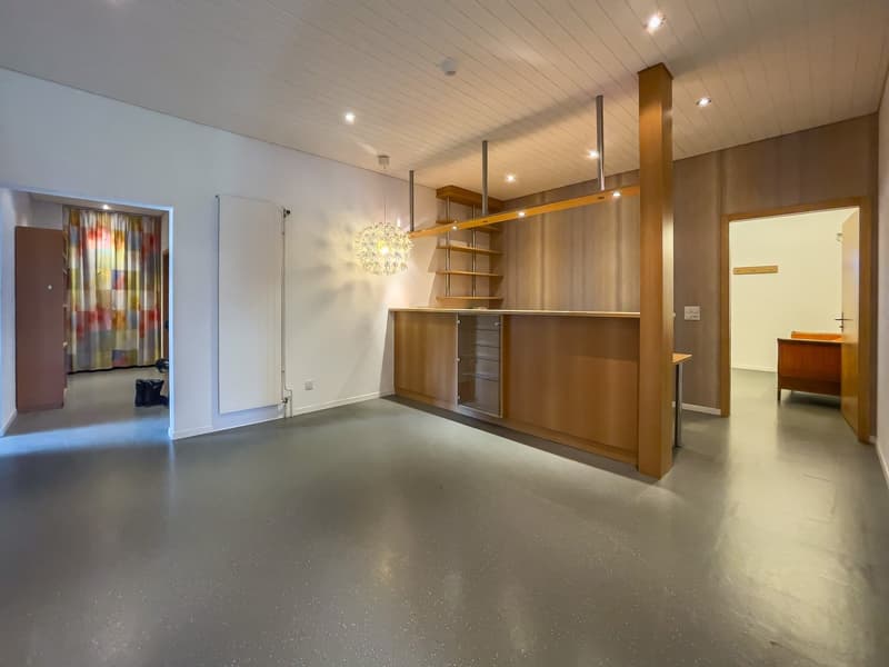 Open-concept living space with wooden shelving unit, modern lighting fixtures, and a view of an adjacent room through an open doorway.
