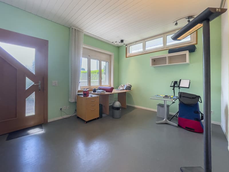 Desk, two drawers, wastebasket, windows, light fixture, floor mat, office chair, table with items
