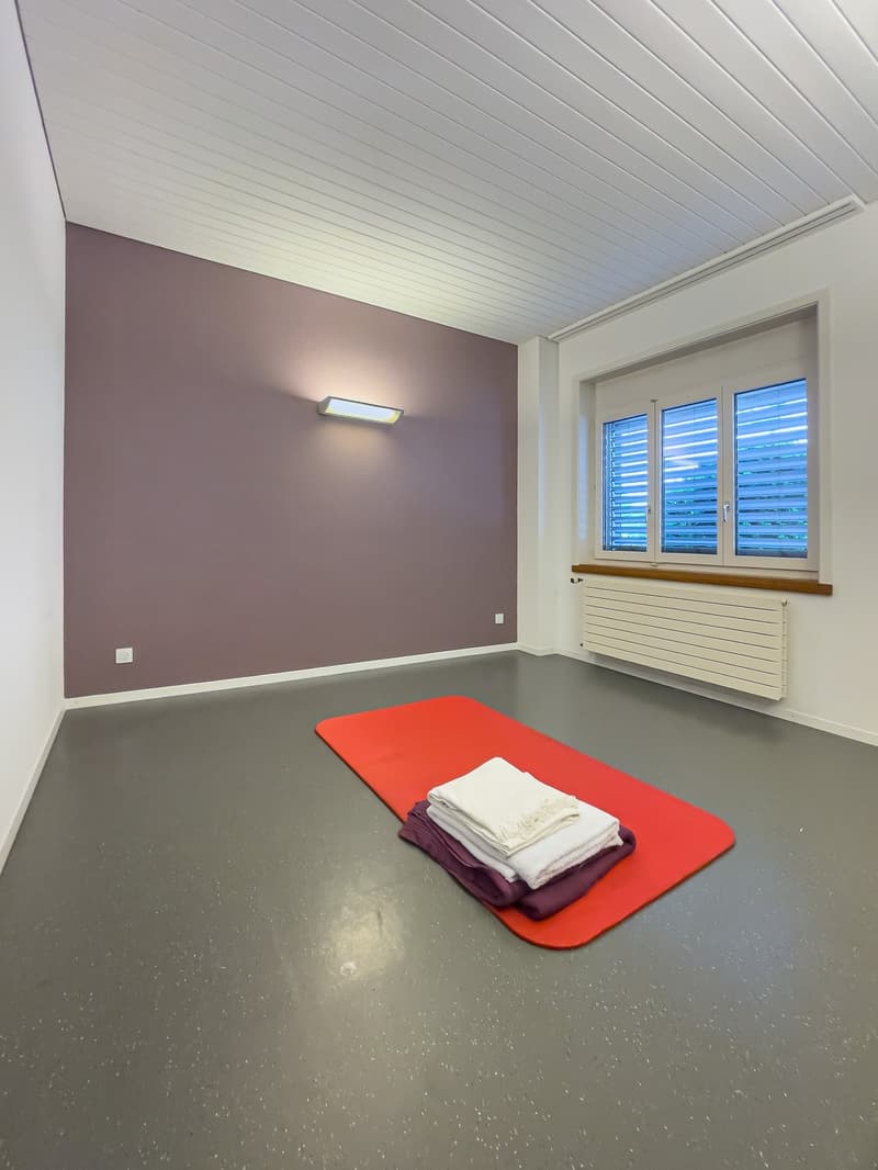 Empty room with gray flooring, a red yoga mat, stacked towels, and a wall-mounted light. There is a white wall radiator beneath a window with closed blinds.