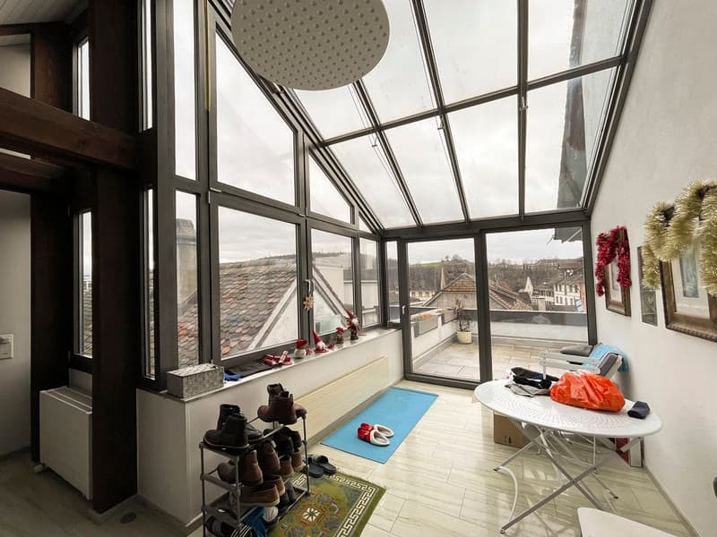 Room with large windows, a white table, a rug, and a shower head on the ceiling. The view outside the windows shows a snowy rooftop and cityscape.