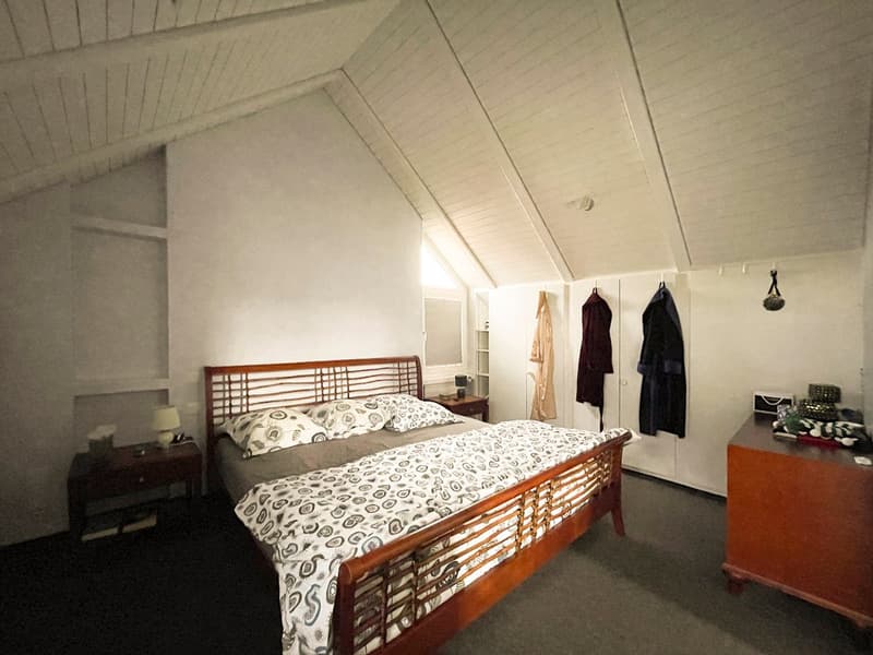 Large wooden bed with wooden headboard, two nightstands, carpeted floor, white walls, ceiling with wooden beams, clothes hanging on the wall, mirror