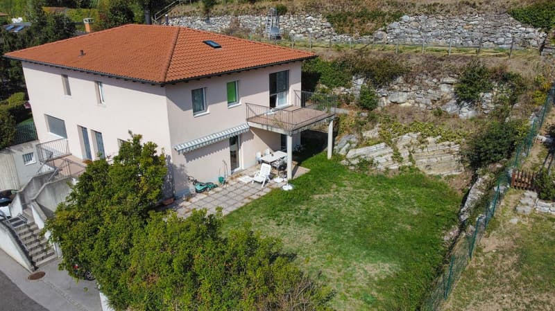 two story house with brown tiled roof, multiple windows, small balcony at ground level, garden, lawn, outdoor table, lawn mower