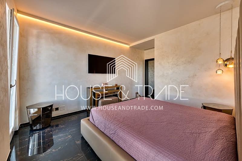 large bed, pink bedspread, white walls, ceiling lighting, modern decor, small table, desk with TV, shiny table