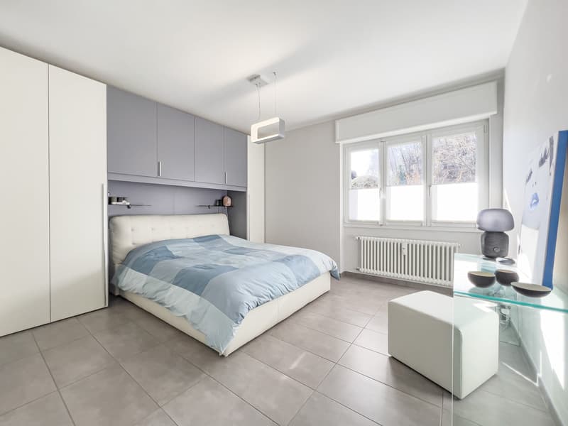 modern bedroom with white bed, white headboard, white wardrobe, white ottoman, gray tiled floor, white walls, large window, radiator, ceiling lamp, glass table, lamp
